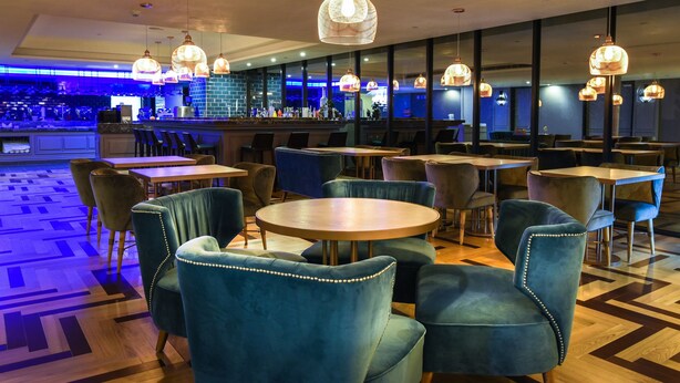 A restaurant with round tables and blue upholstered chairs, a bar area with a blue-lit counter, and hanging pendant lights.
