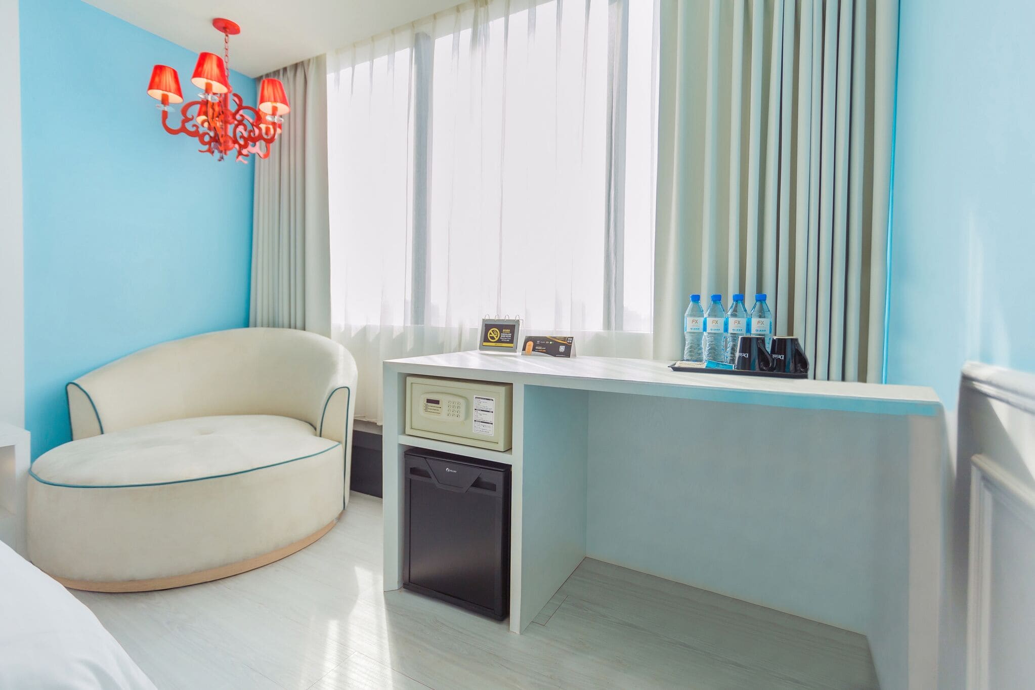 A compact hotel room with a light blue wall, a white desk with a small refrigerator, a cream-colored armchair, a red chandelier, and a window with curtains.