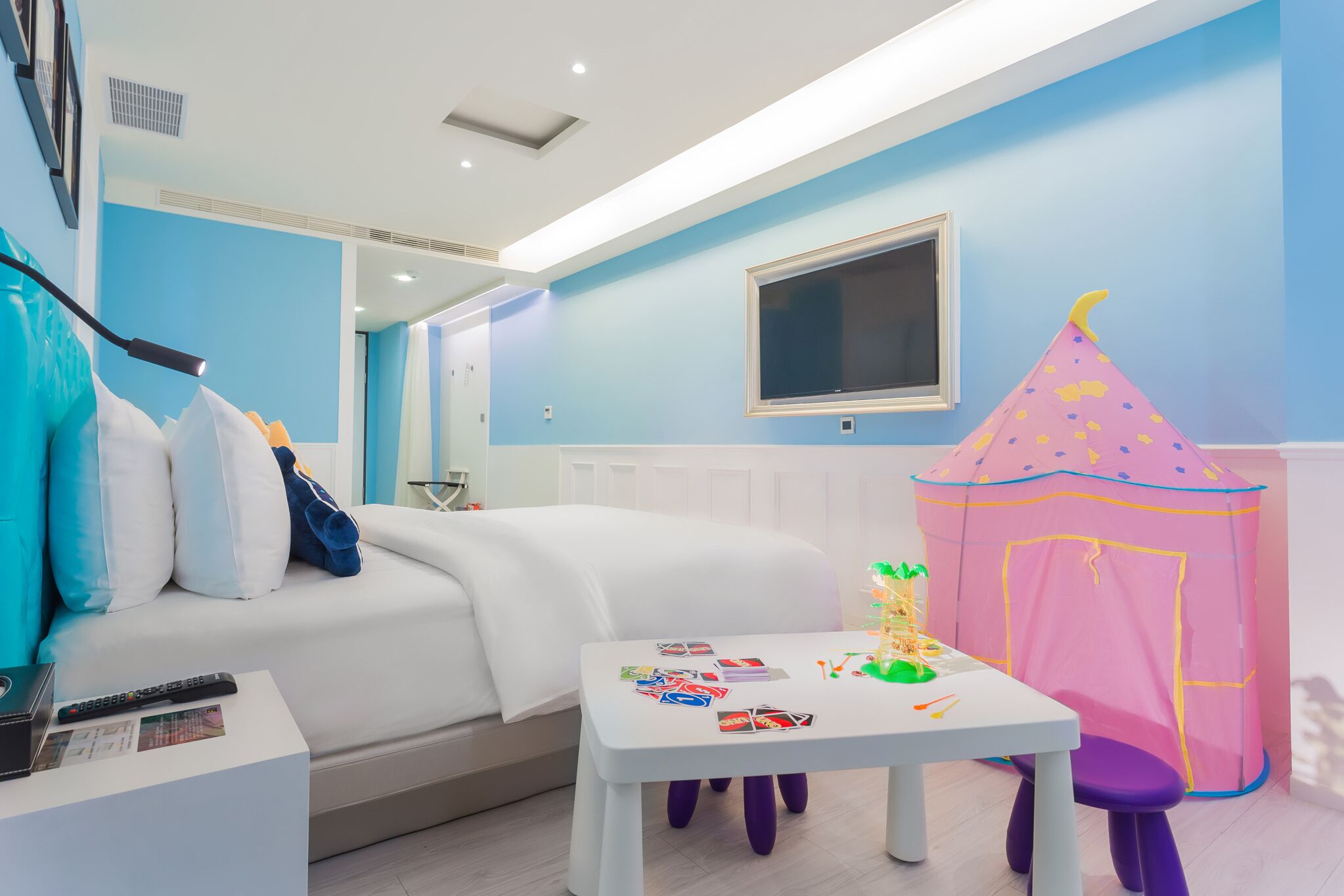 A bedroom with a bed, a small table with toys, a pink play tent, and a TV on the wall.
