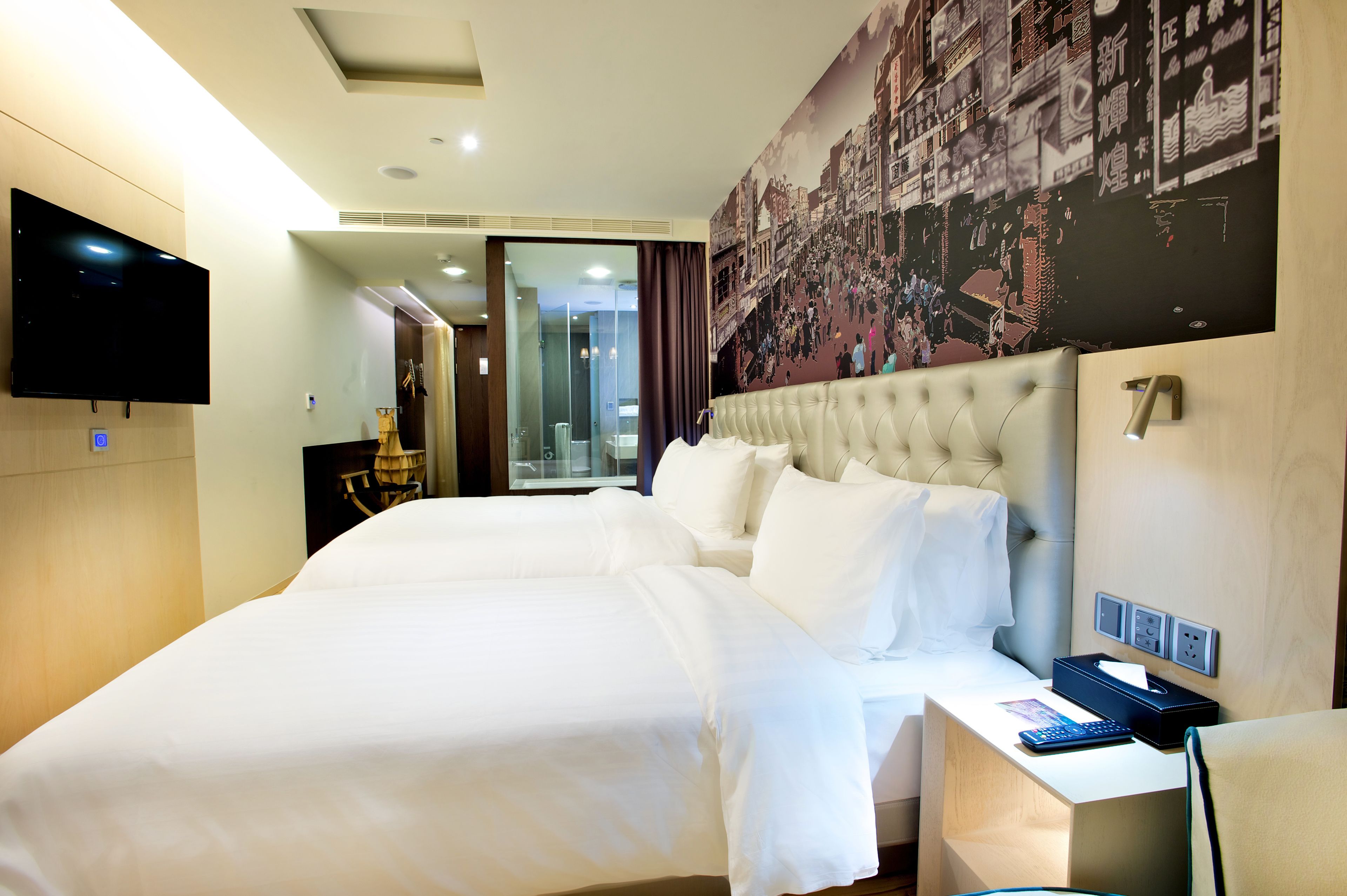 A hotel room with two beds, a flat-screen TV, a desk with a computer, and a large cityscape mural on the wall.