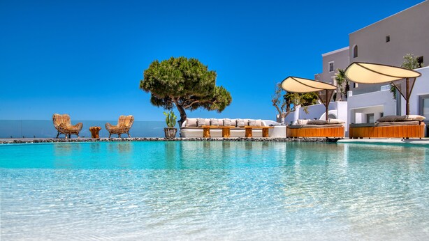 A pool area with lounge chairs, a sofa, and umbrellas, surrounded by a white building with a clear blue sky in the background. - San Marino Suites by Calm Collection (Santorini)
