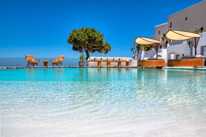 A pool area with lounge chairs, a sofa, and umbrellas, surrounded by a white building with a clear blue sky in the background. - San Marino Suites by Calm Collection (Santorini)