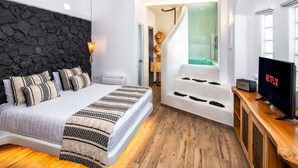 A modern bedroom with a stone wall, a large bed with patterned bedding, a wooden floor, a TV, and a bathroom with a bathtub and shower. - San Marino Suites by Calm Collection (Santorini)