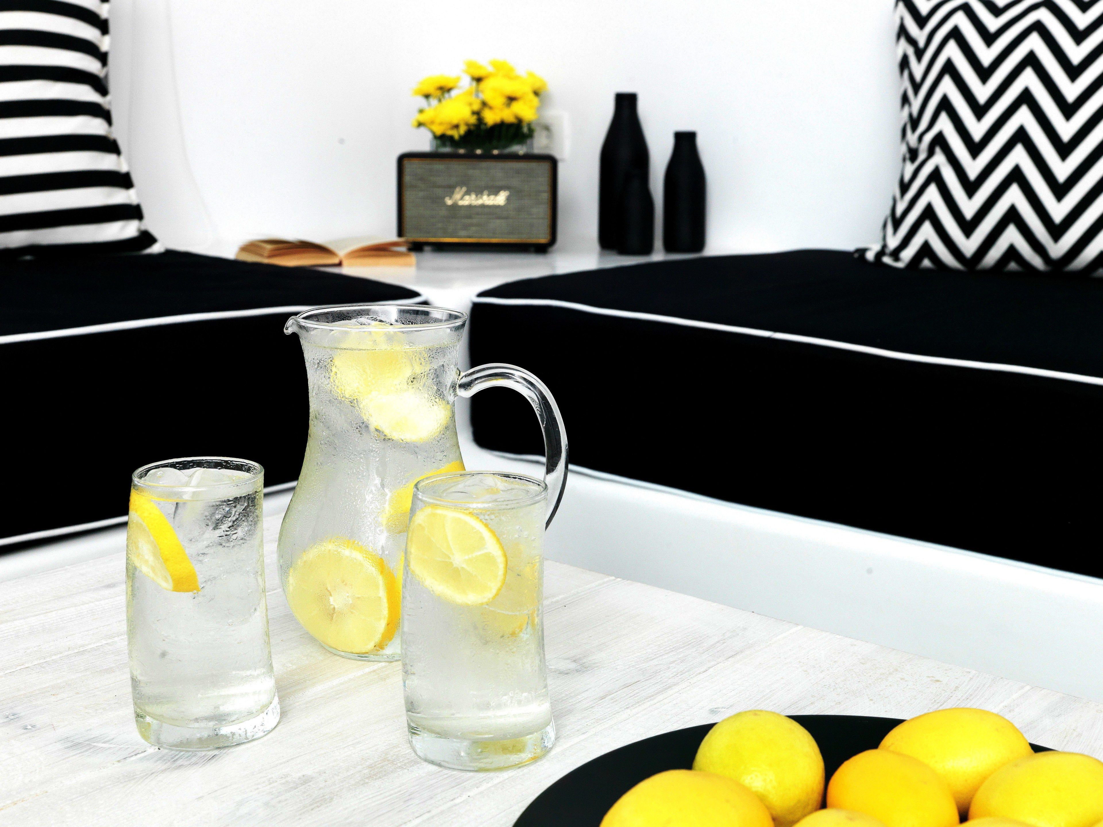 A clear pitcher and two glasses of lemonade with lemon slices, a black and white patterned pillow, a small vase with yellow flowers, and a black and white patterned cushion on a white table.