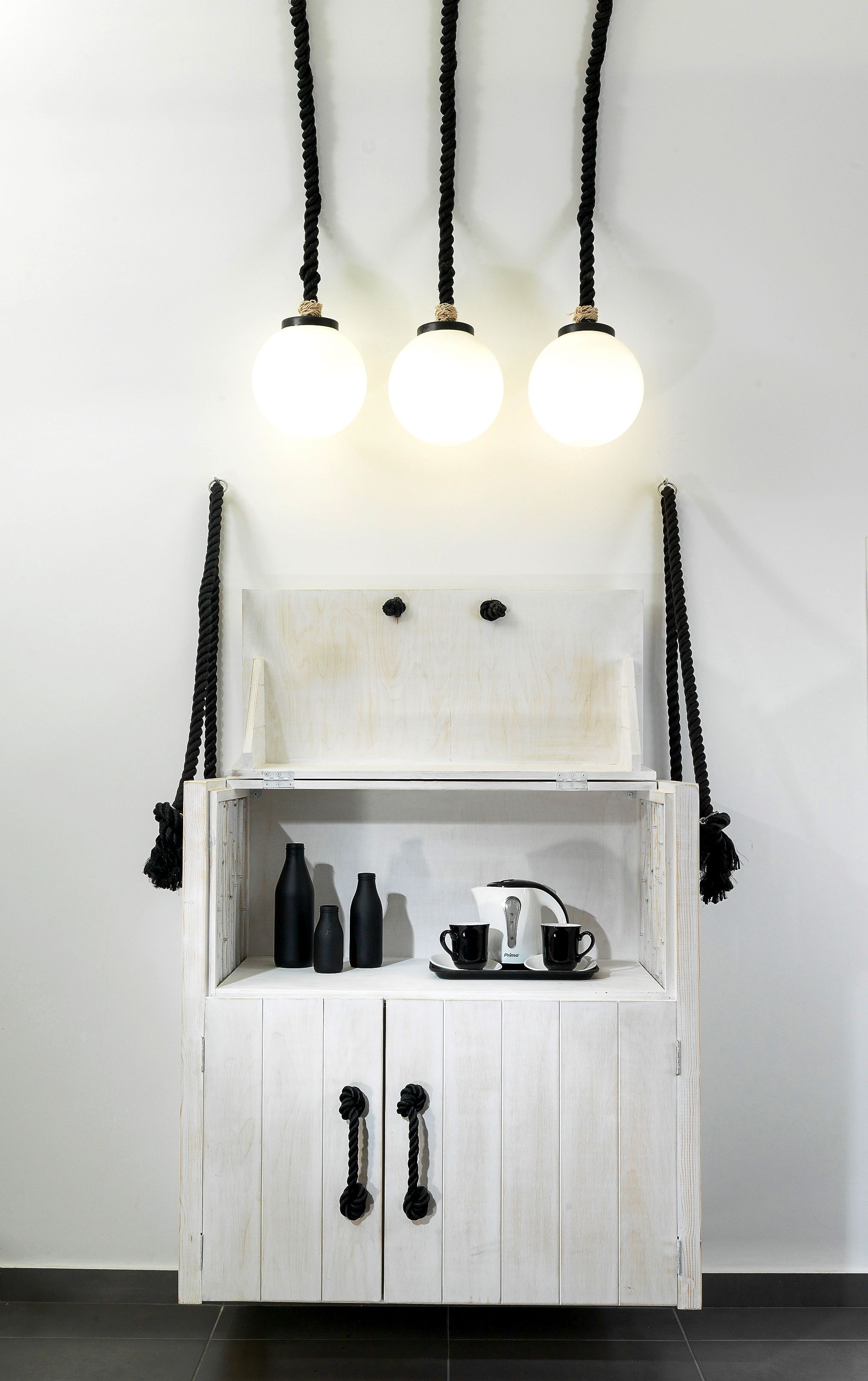 Un meuble blanc en bois, doté de poignées en corde noire, sur lequel sont posés une machine à café et des tasses noires.