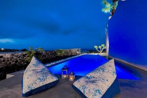 A poolside area with two seating cushions, a stone wall, and a view of the ocean. - San Marino Suites by Calm Collection (Santorini)