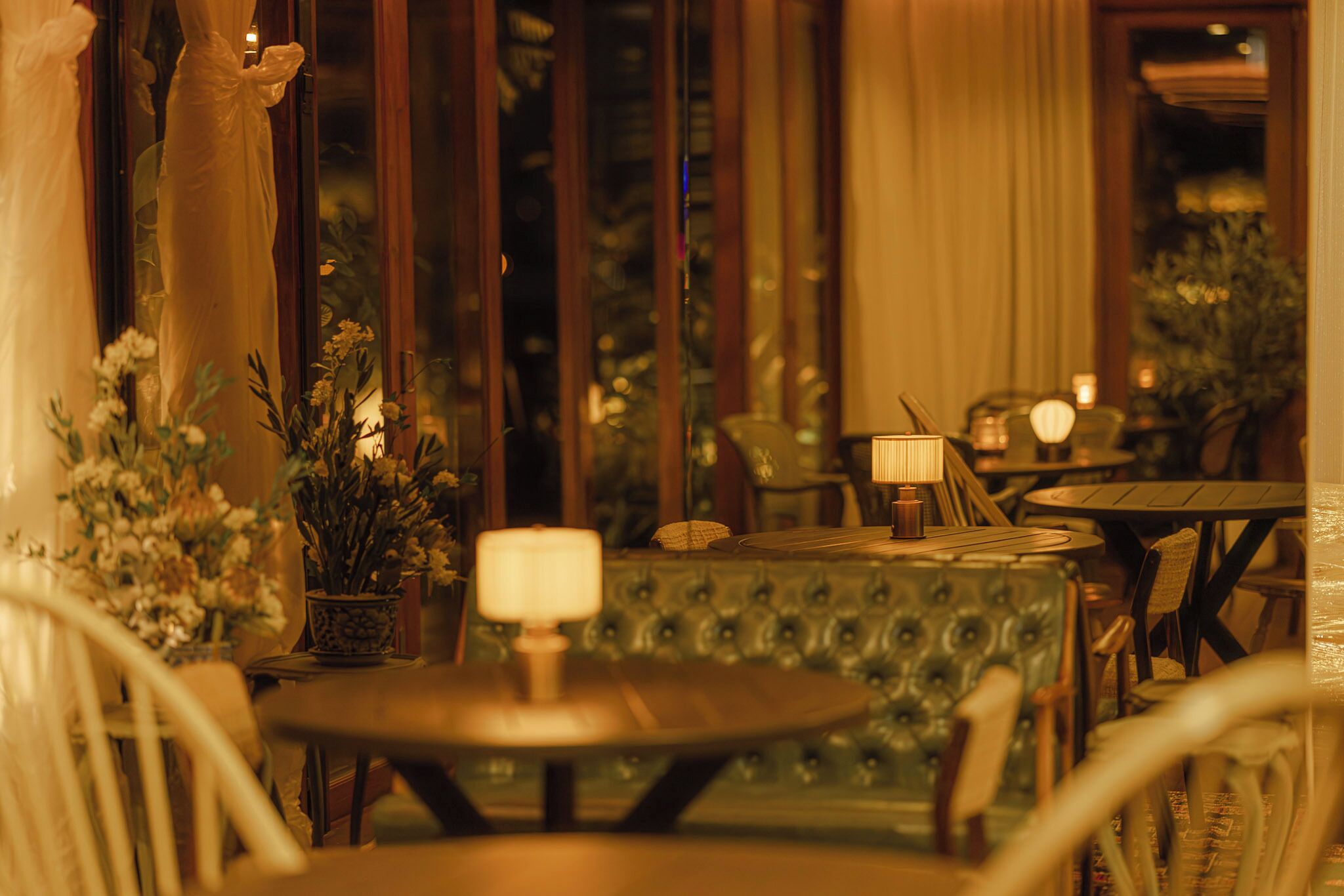 A cozy indoor seating area with plush chairs and tables, a decorative lamp, and a view through a window with curtains.