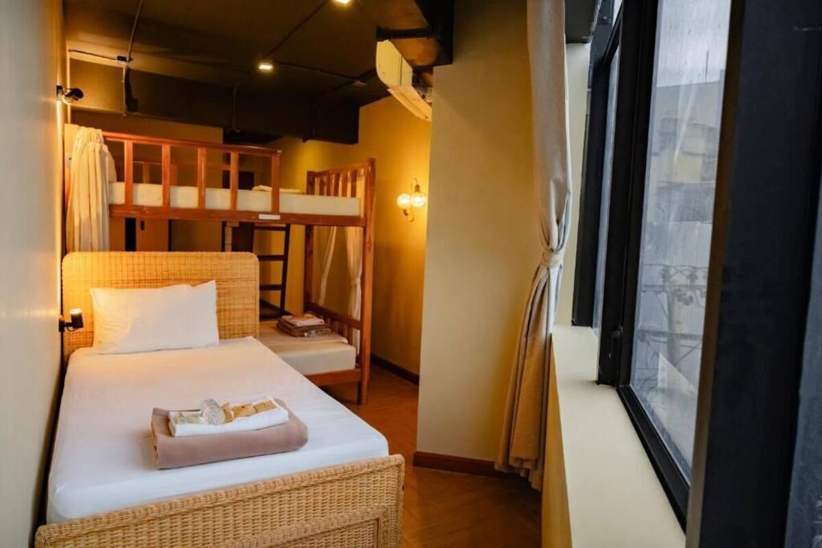 Una cama individual con cabecera, una cama nido inferior, una silla de mimbre y una ventana con cortinas.
