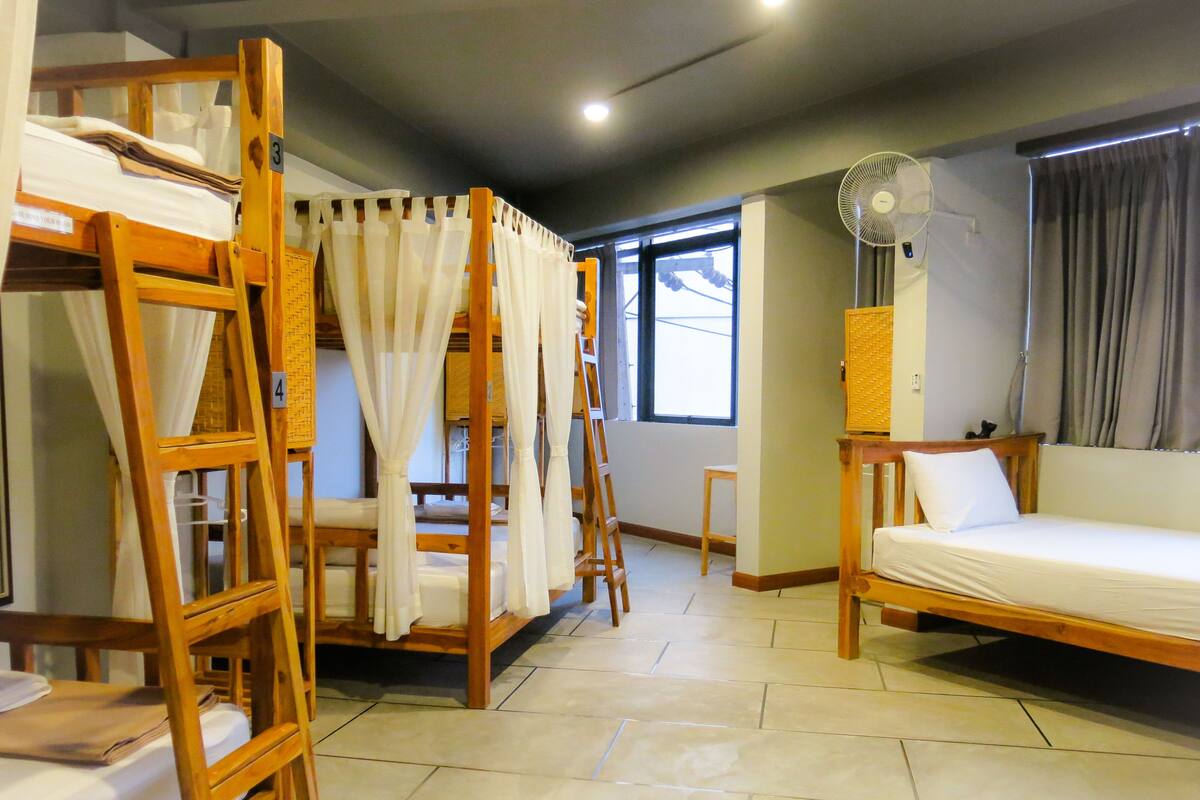 Una habitación con literas, una cama individual, un ventilador de pie y una ventana con cortinas.