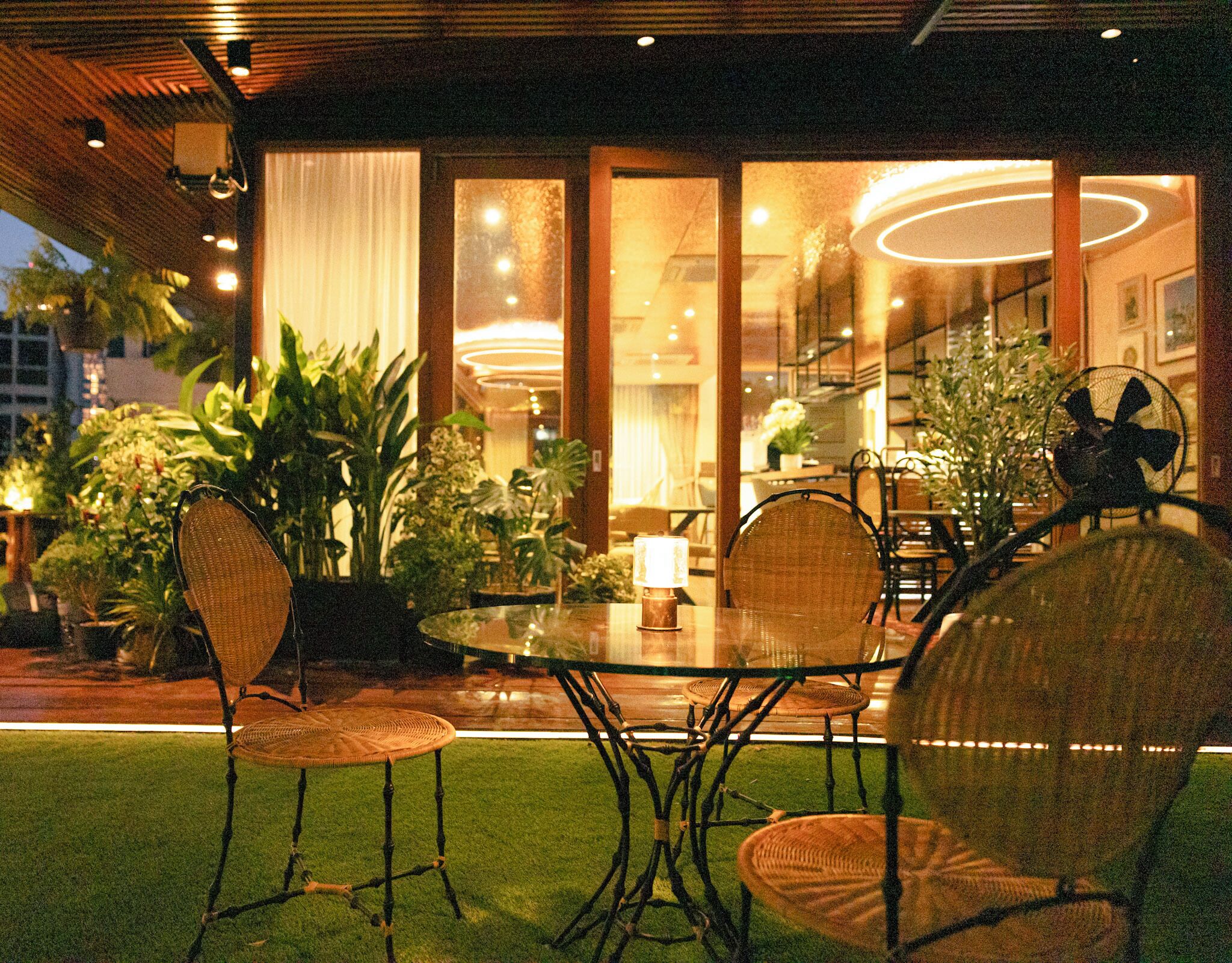 An outdoor dining area with wicker chairs and a glass table, surrounded by potted plants and a view of the interior.