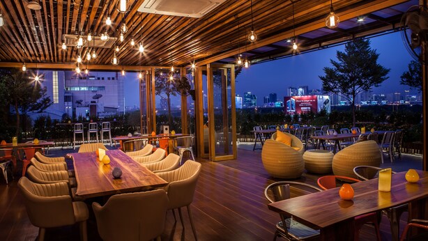 An outdoor dining area with wooden tables and chairs, large windows, and a city view at dusk.