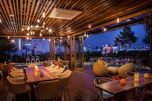 Un espace de restauration en plein air avec des tables et des chaises en bois, de grandes fenêtres et une vue sur la ville au crépuscule.