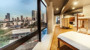 A modern hotel room with a large window offering a city view, a bunk bed, a desk, and a rattan room divider.