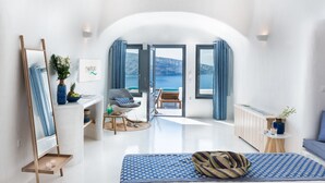 Une chambre moderne et bien éclairée, dotée d’un grand lit, d’un coin salon avec un fauteuil et une table, d’un miroir et offrant une vue sur la mer grâce à des portes ouvertes.