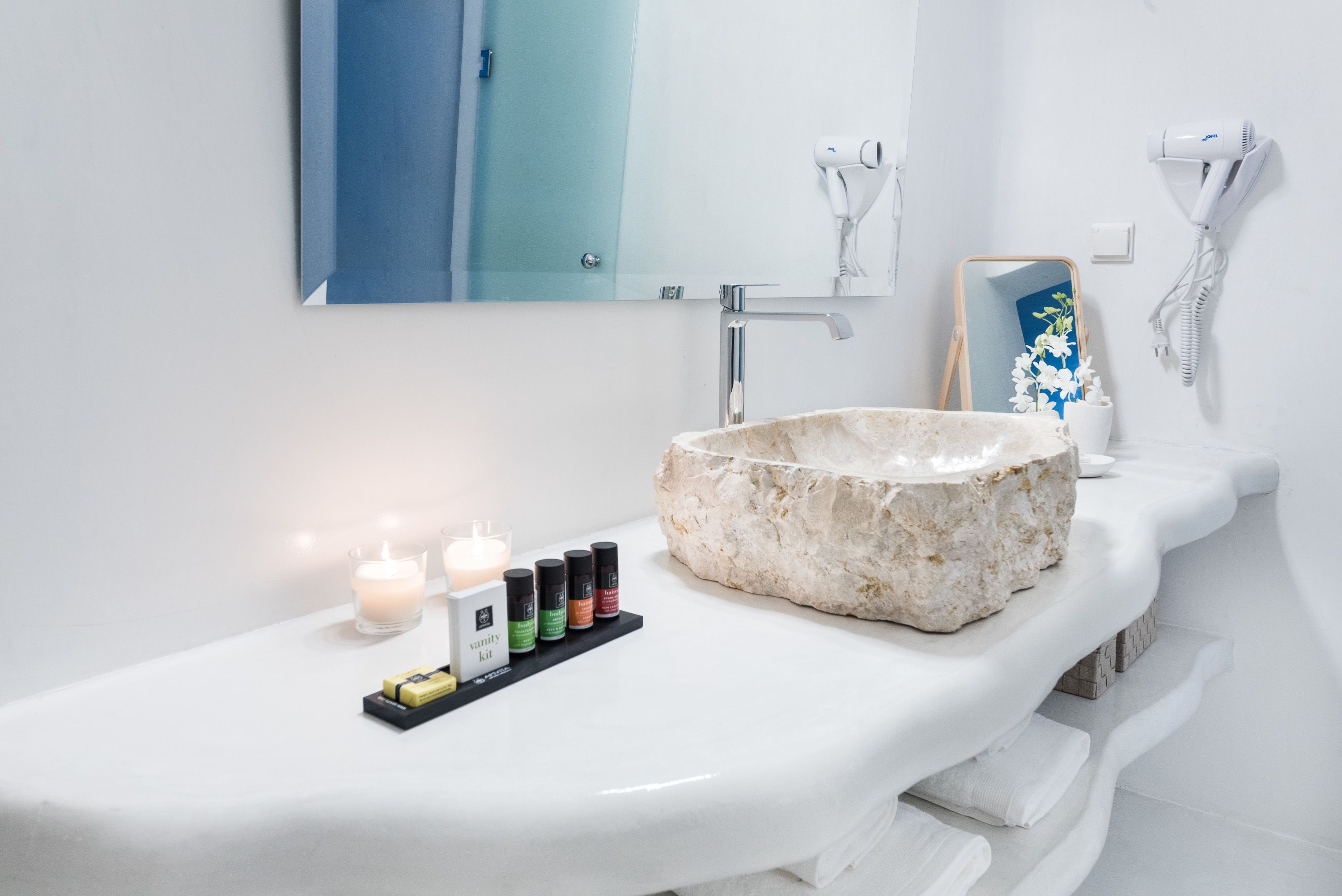 A bathroom with a white sink, a large stone basin, a mirror, and a vanity with a shelf holding candles and a vanity kit.