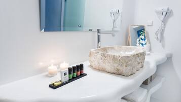 A bathroom with a white sink, a large stone basin, a mirror, and a vanity with a shelf holding candles and a vanity kit.