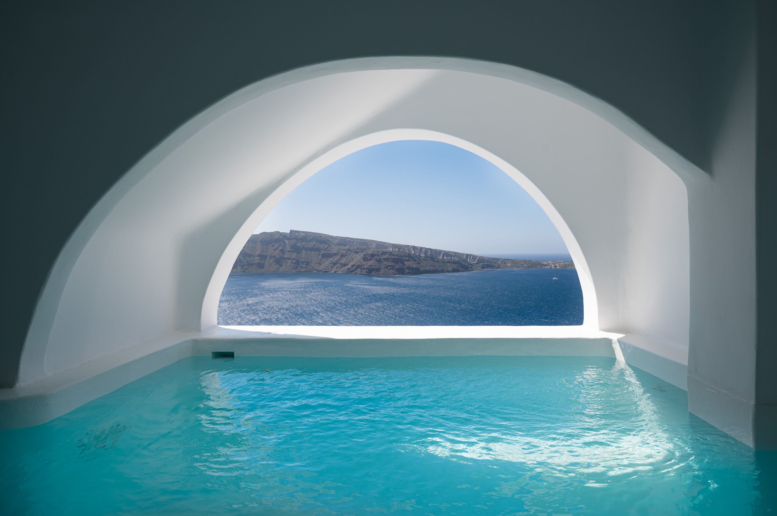 A room with a large arched window overlooking a clear blue sea and a rocky coastline.