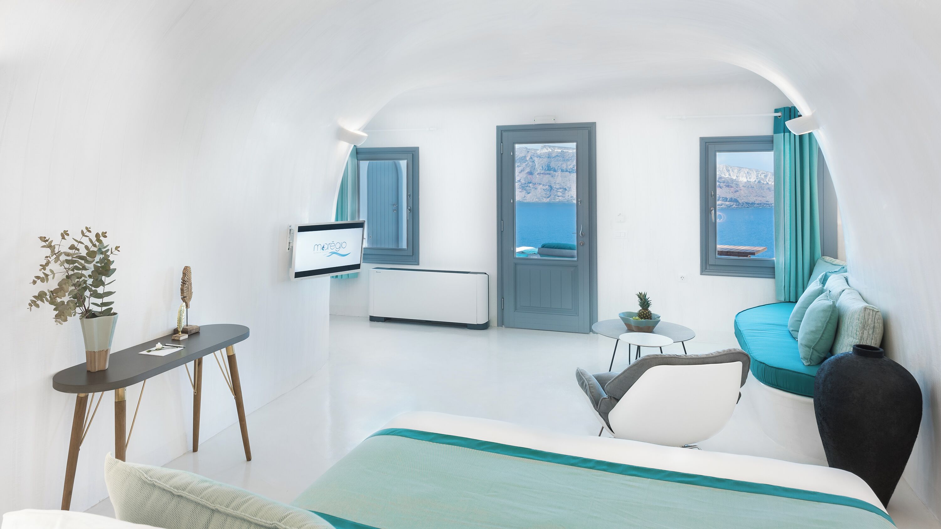 Una habitación moderna y minimalista con cama, una mesita, una silla y televisión. Hay dos ventanas con cortinas, y se ve el mar a través de ellas.