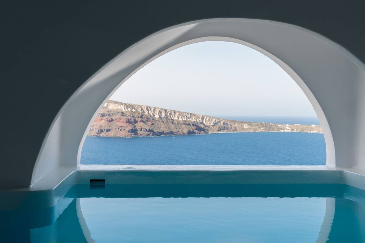 Vista desde un ventanal de arco que muestra una zona de piscina con vistas a un paisaje costero con acantilados y el mar.