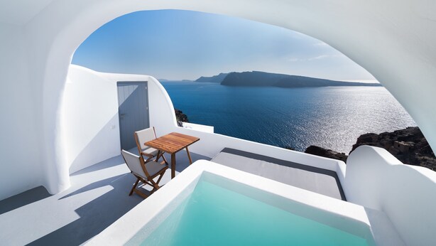 A white-walled infinity pool with a wooden table and chairs, overlooking a clear blue sea and rocky coastline. - Maregio Suites (Santorini)