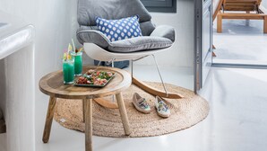 A modern room with a view of the sea, a grey armchair with a blue pillow, a small round table with drinks and shoes, and a large window with curtains. - Maregio Suites (Santorini)