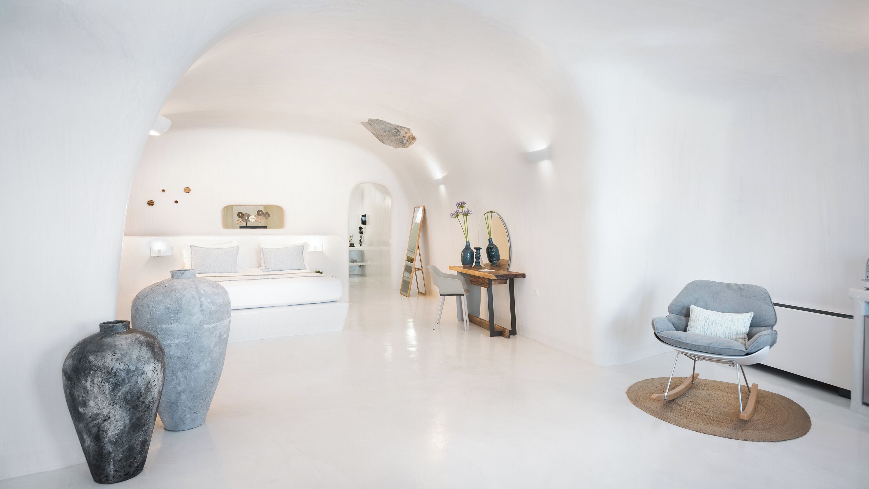 A modern, minimalist room with a large bed, a desk with a chair, and two large vases.