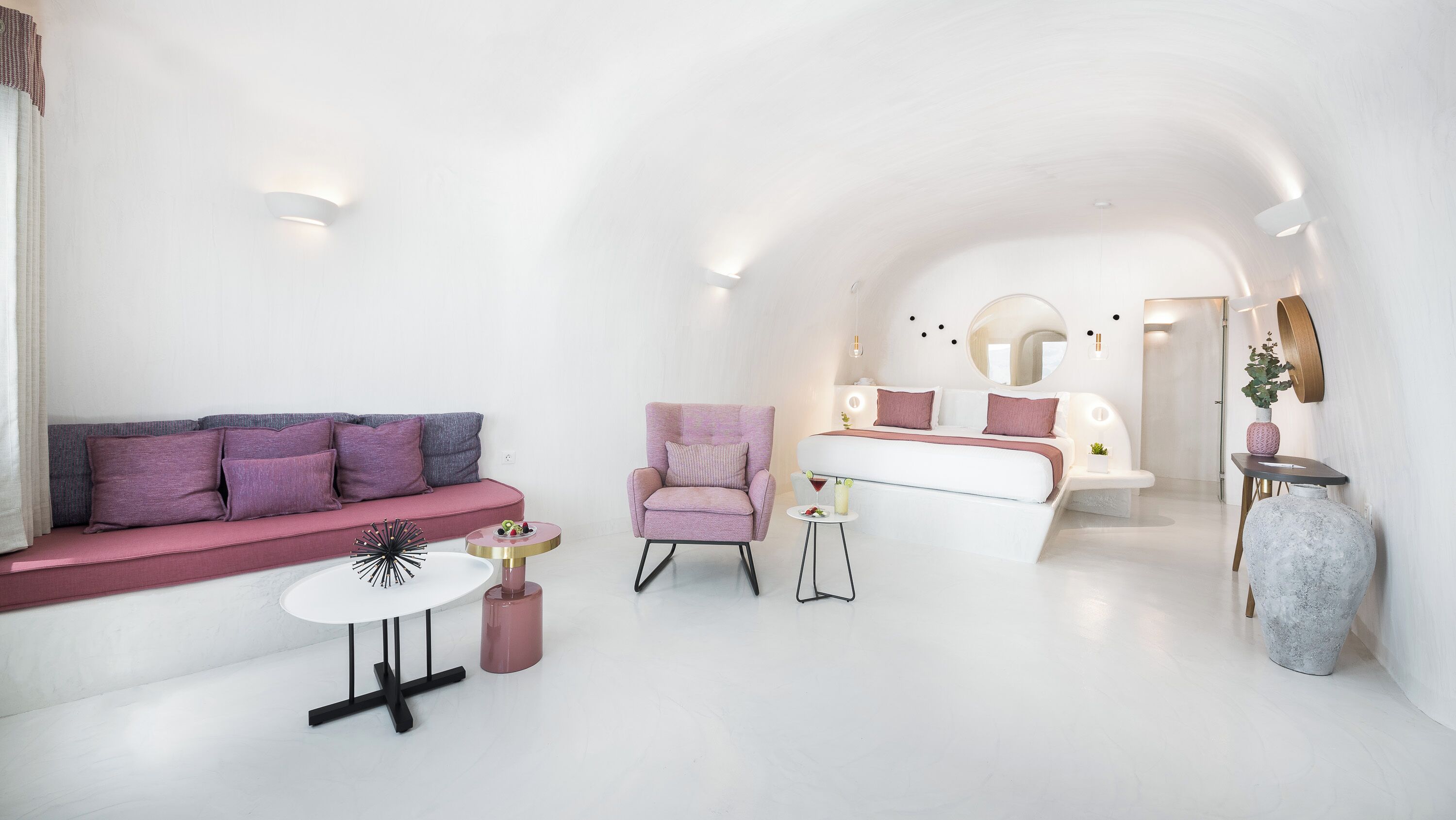 Una habitación moderna y minimalista con un sofá blanco, un sillón rosa, una mesita redonda pequeña y una cama con cabecero blanco.