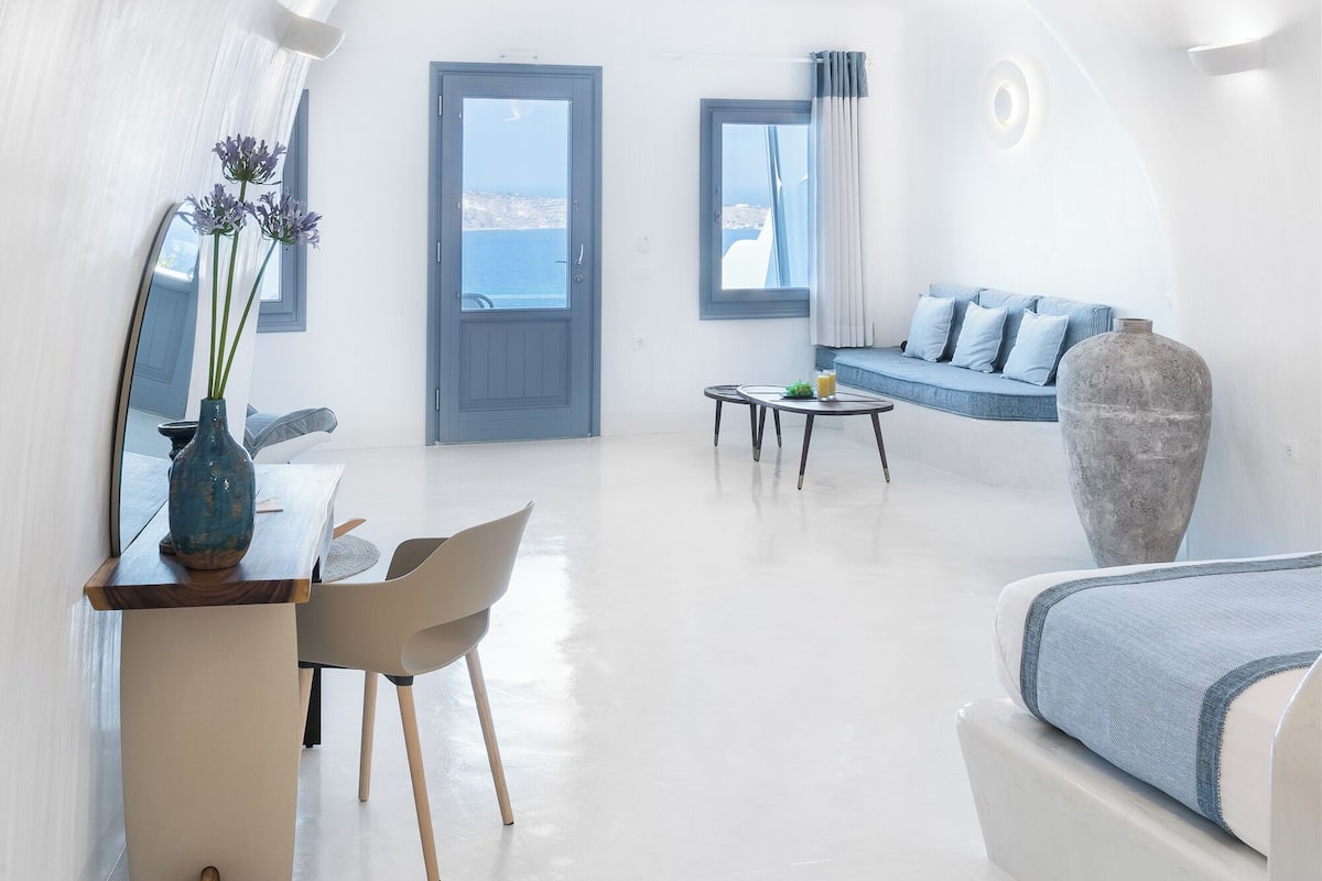 Un interior moderno y blanco con zona de comedor, sofá y grandes ventanales con vistas al mar.