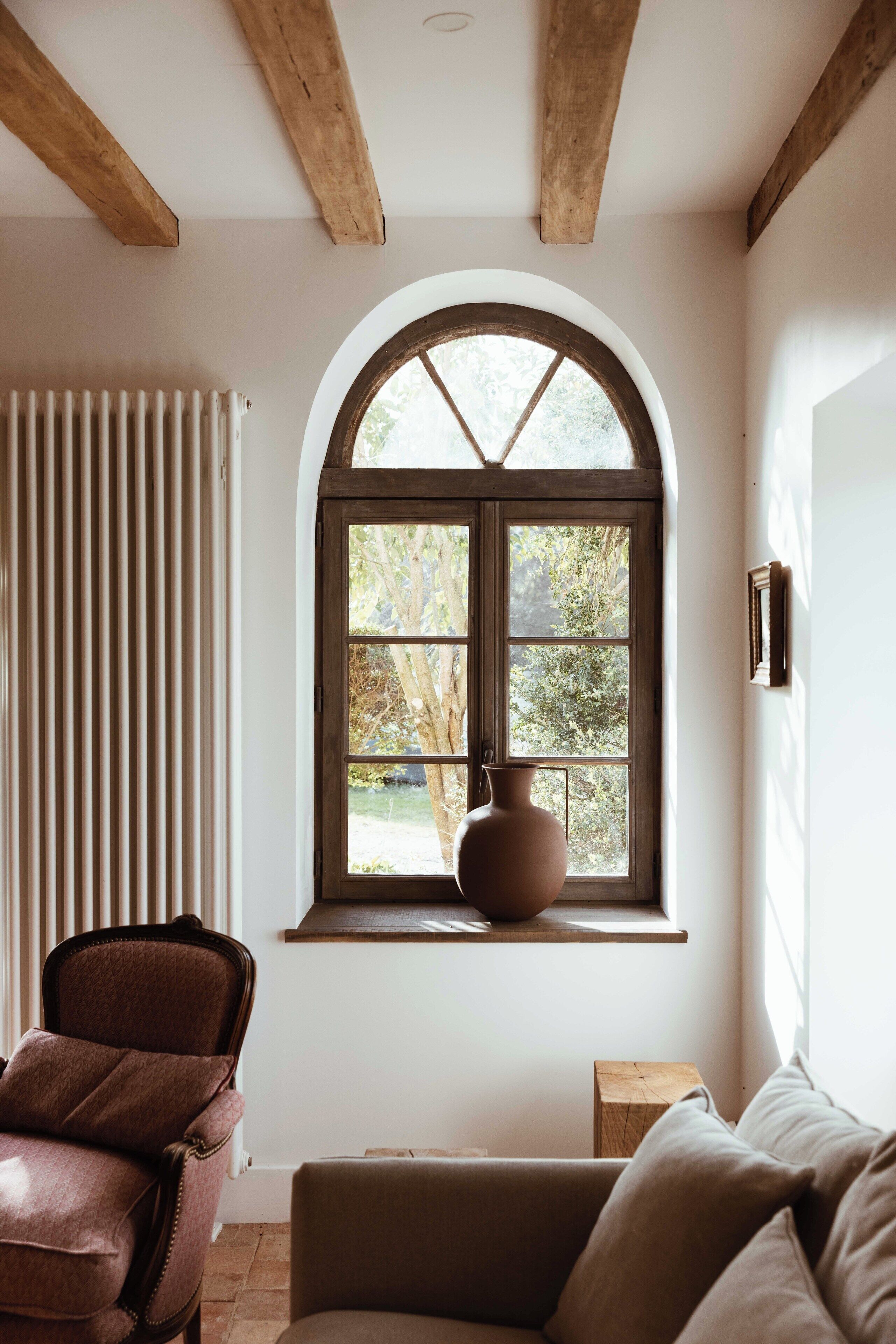 Una habitación con un ventanal arqueado grande, un radiador, un florero marrón sobre una estantería, un sillón de cuero marrón y un sofá.
