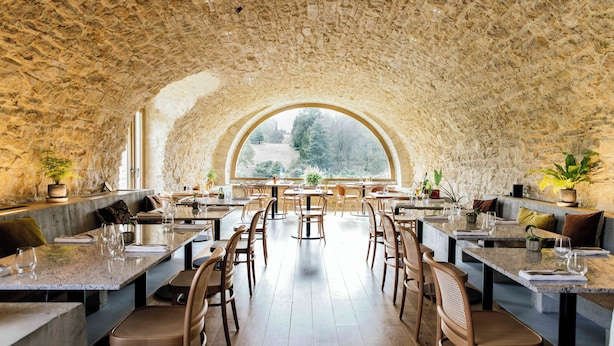 Un restaurante amplio con paredes de piedra altas y un gran ventanal arqueado que da a una zona boscosa. Las mesas están dispuestas con sillas, y hay plantas en macetas sobre ellas.