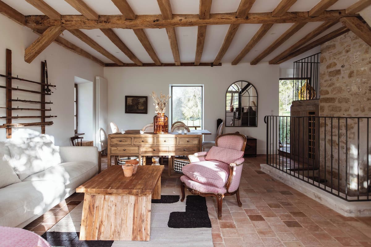 A rustic interior with exposed wooden beams, a stone wall, a white sofa, a wooden coffee table, and a pink armchair.