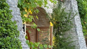 Un rústico edificio de piedra con puerta de madera, rodeado de abundante vegetación y una terraza de madera.