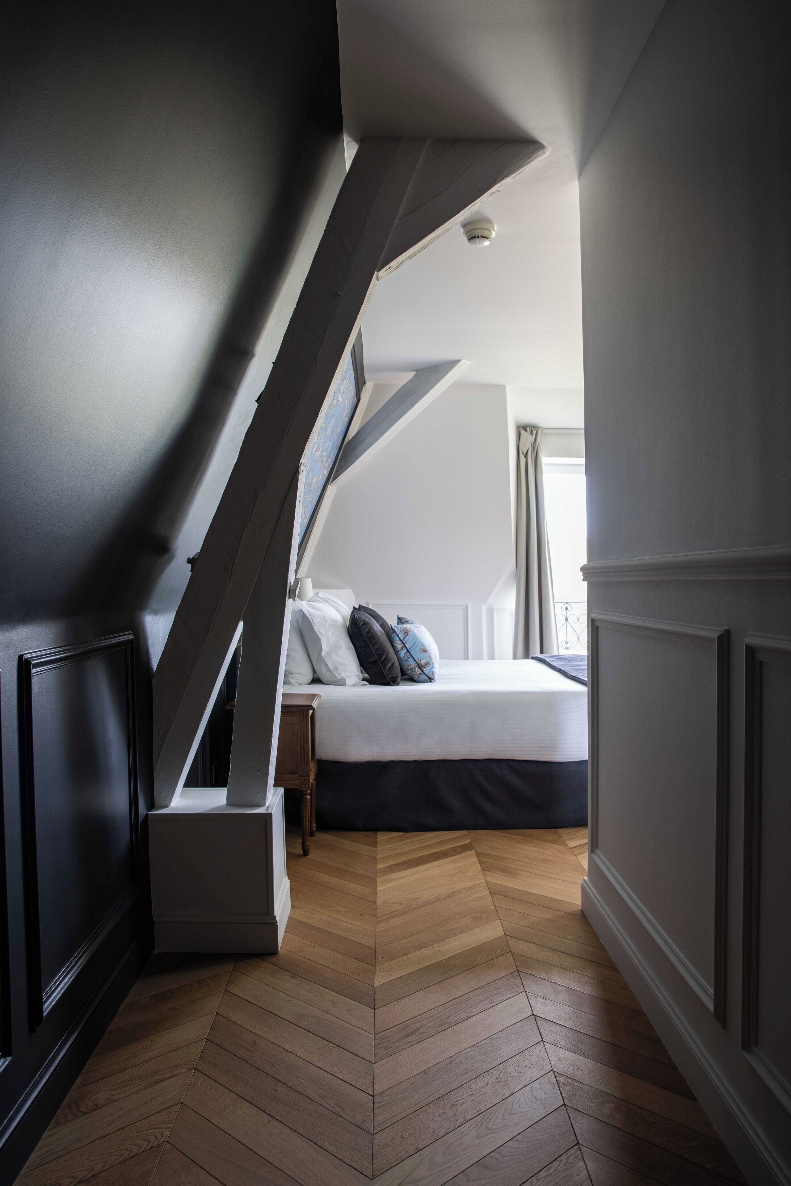 A bedroom with a herringbone-patterned wooden floor, a bed with white bedding and a dark headboard, and a window with curtains.