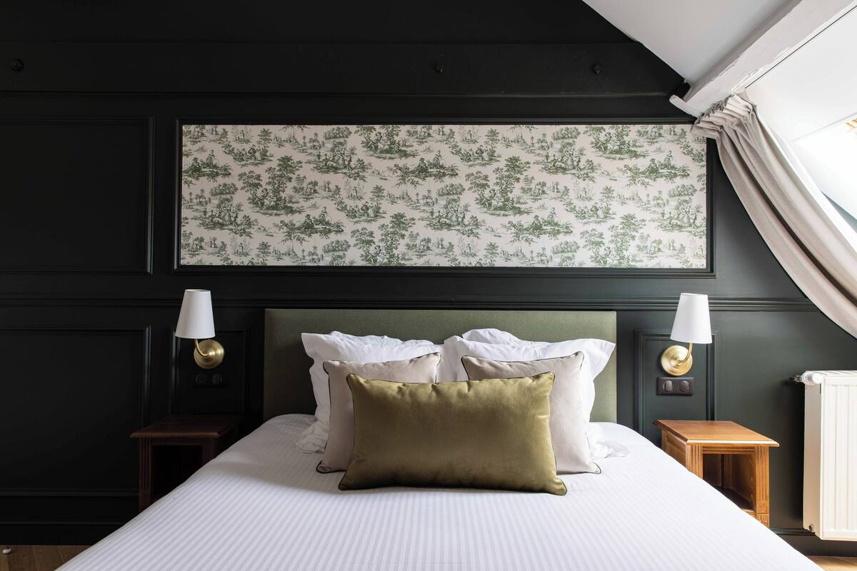 A neatly made bed with white linens and a green pillow, flanked by two bedside tables with lamps, in a room with dark walls and a large botanical wallpaper.