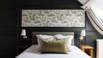 A neatly made bed with white linens and a green pillow, flanked by two bedside tables with lamps, in a room with dark walls and a large botanical wallpaper.