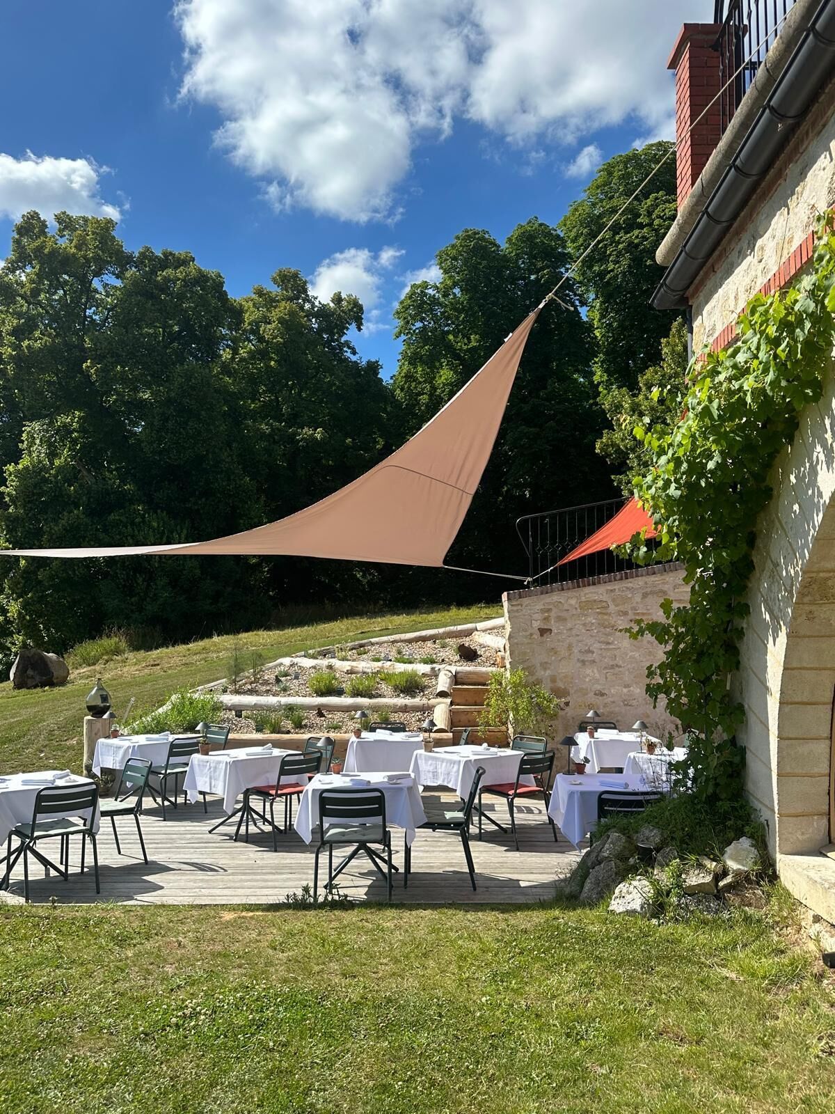 Un'area pranzo all'aperto con tavoli e sedie, una grande vela parasole e un muro in pietra.