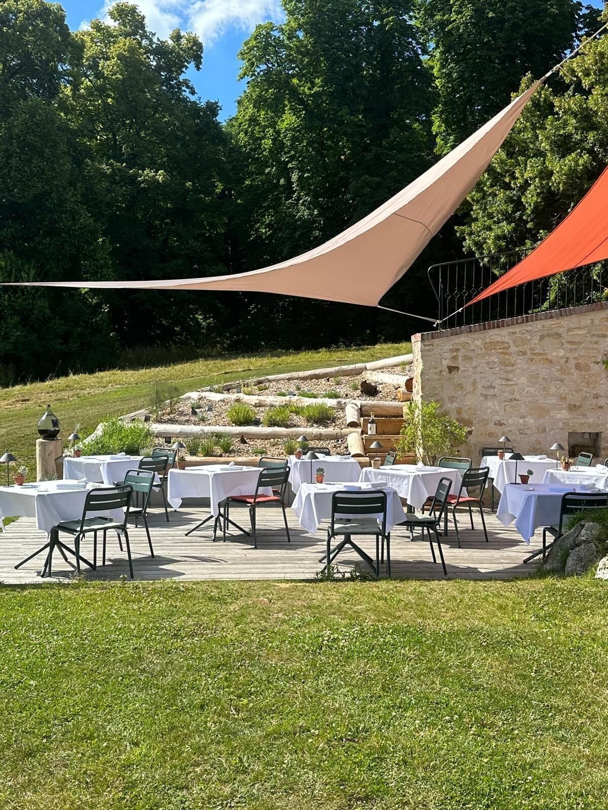 An outdoor dining area with tables and chairs, surrounded by greenery and stone walls.