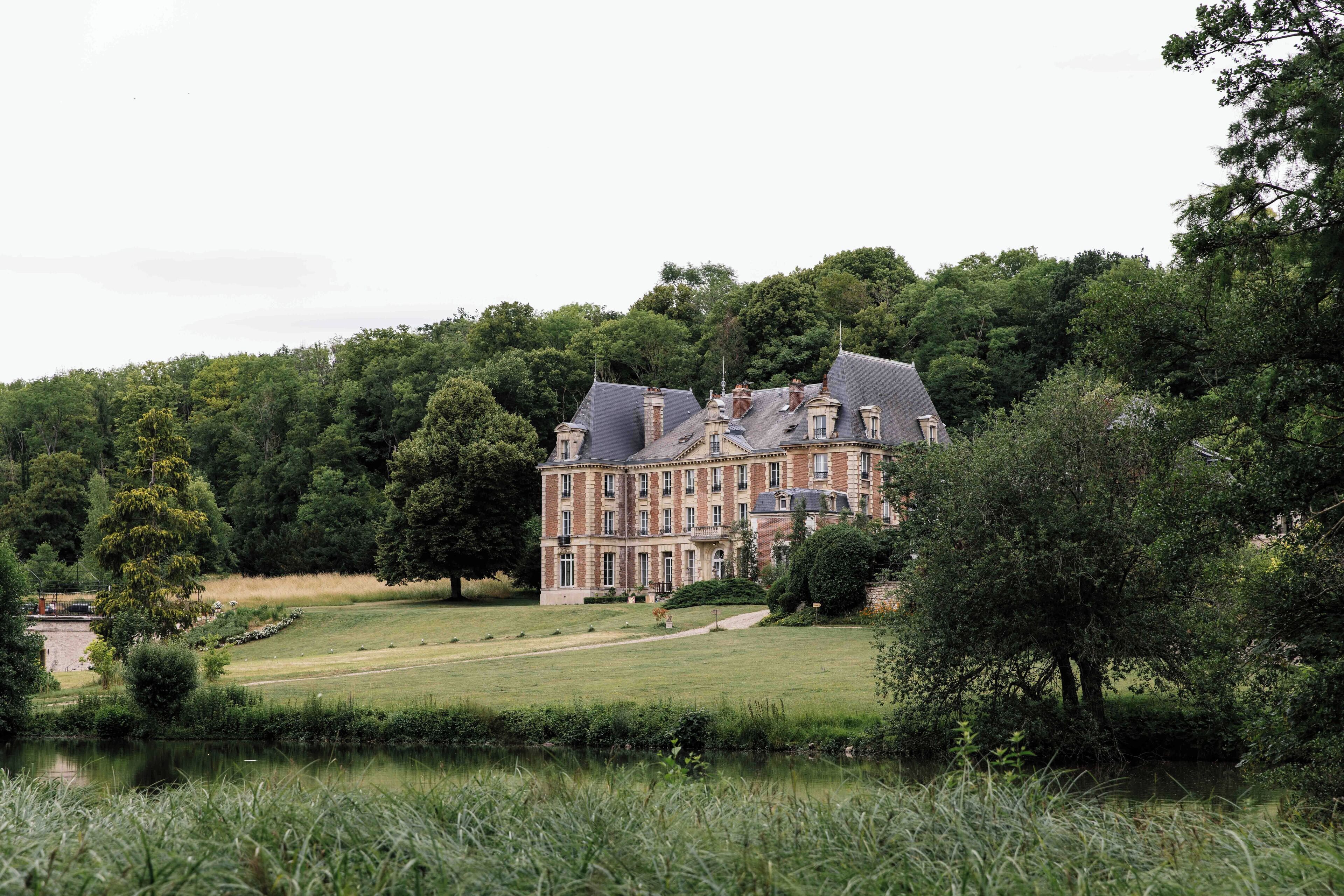 A large, historic building with multiple stories and a prominent roofline, surrounded by lush greenery and a body of water.