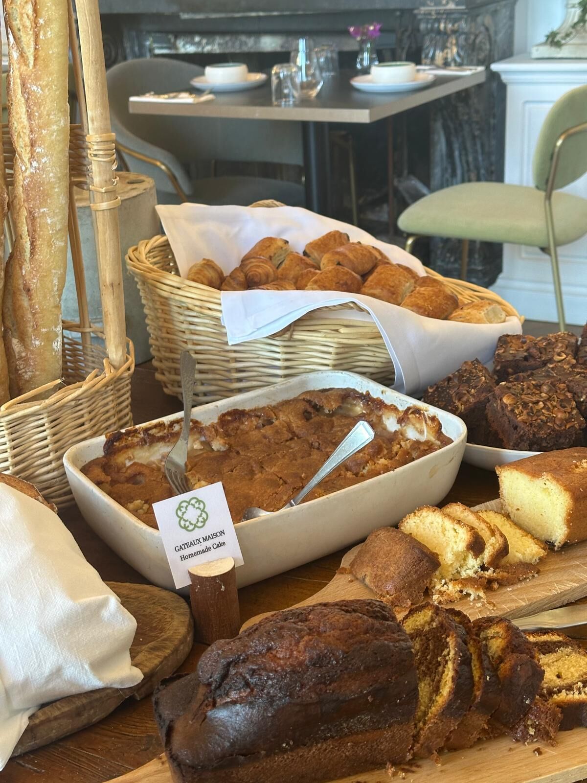 A variety of baked goods including bread, croissants, and cakes displayed on a table in a dining area with a marble countertop and a chair.