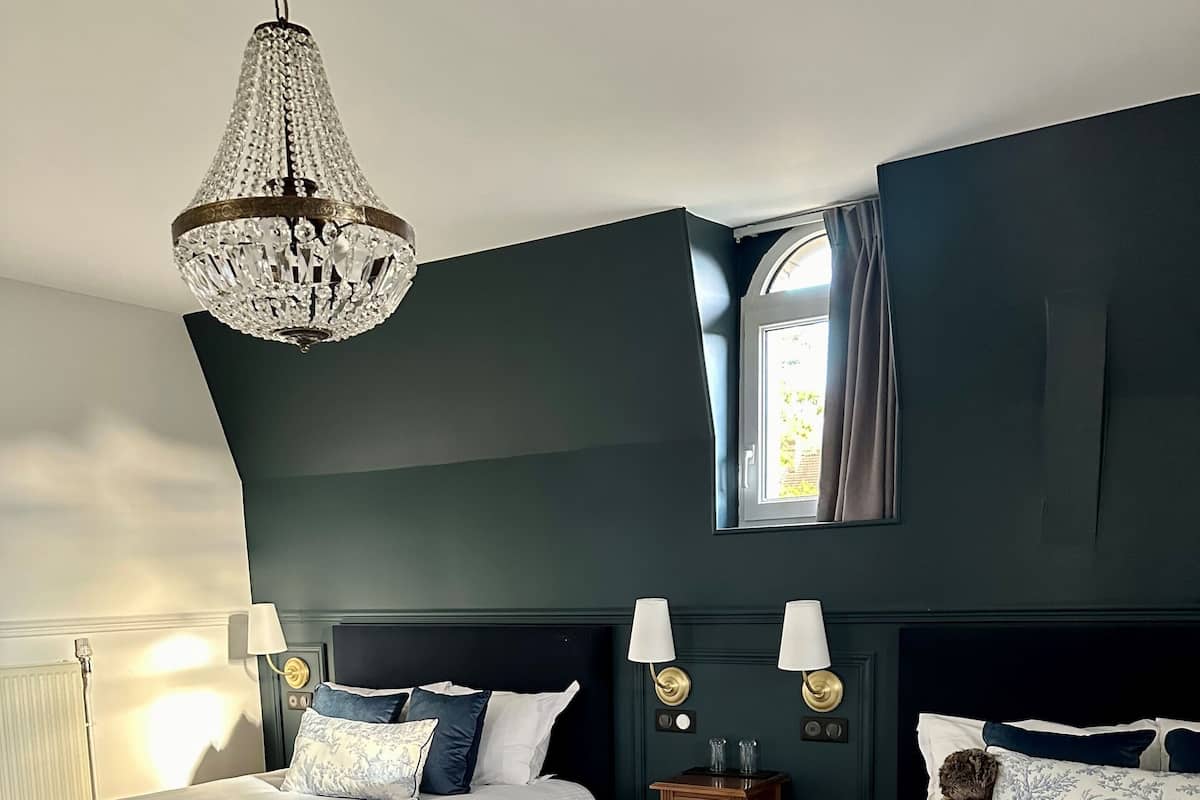 A bedroom with two beds, dark walls, a chandelier, and a window with curtains.