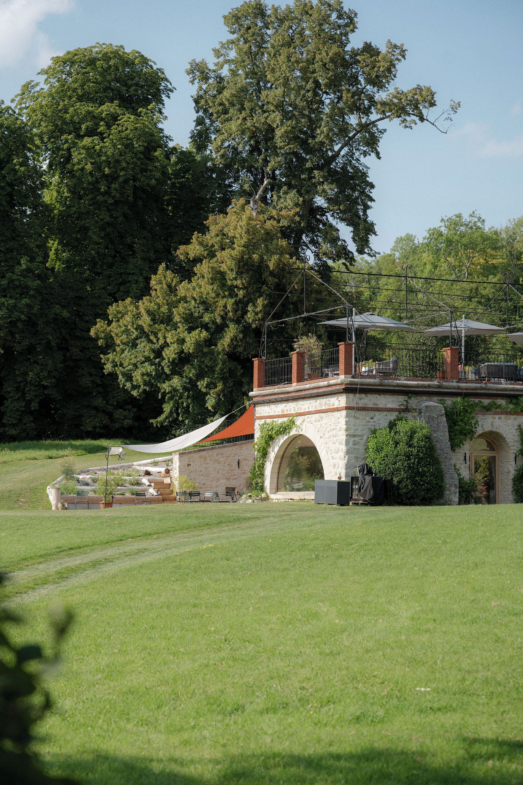 Un bâtiment en pierre doté d’une entrée en arc de cercle, d’un patio couvert aménagé avec du mobilier d’extérieur et d’un grand arbre en arrière-plan.
