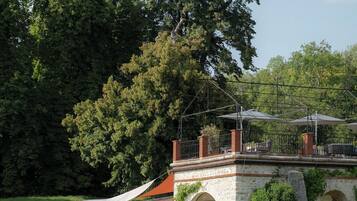 Un bâtiment en pierre doté d’une entrée en arc de cercle, d’un patio couvert aménagé avec du mobilier d’extérieur et d’un grand arbre en arrière-plan.