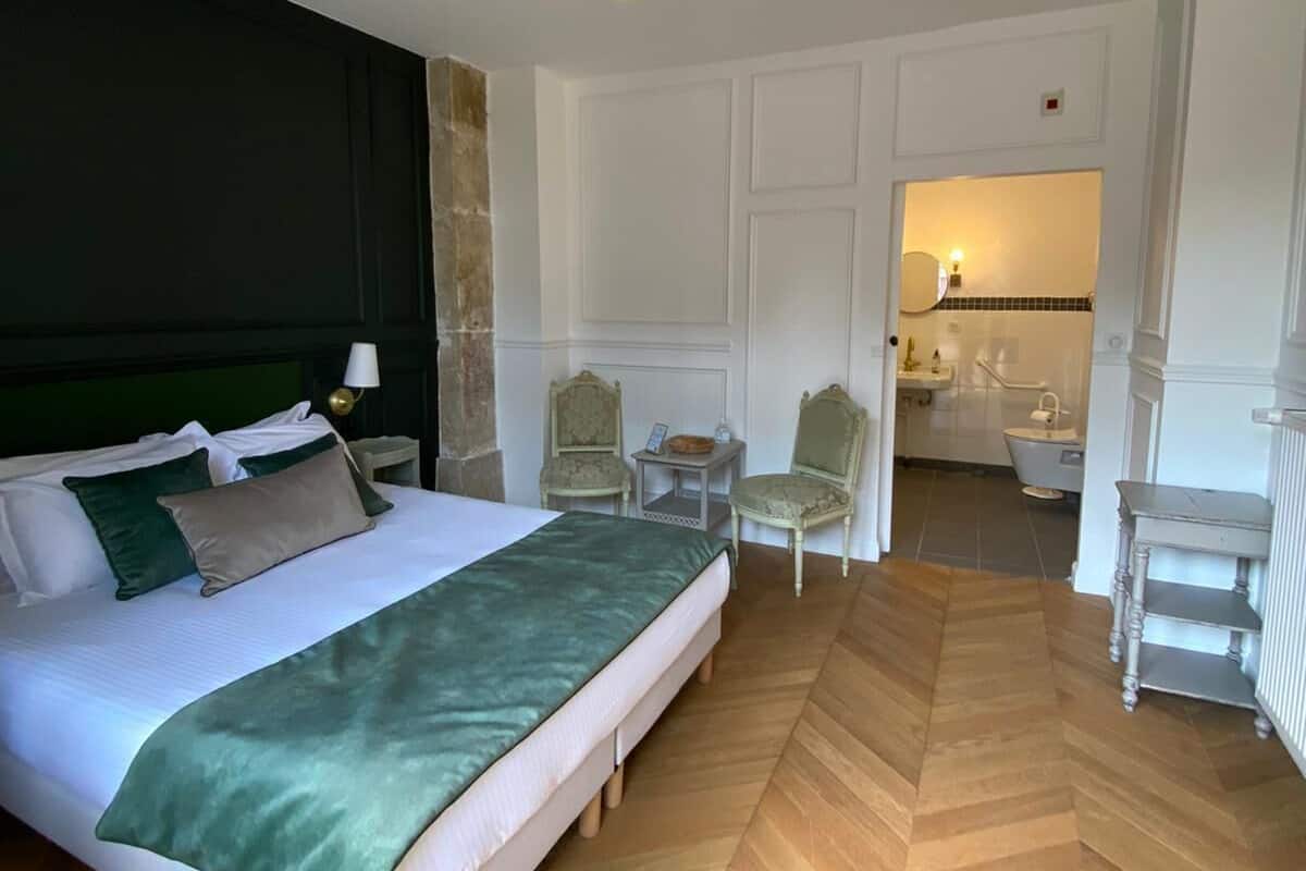 A hotel room with a large bed, two chairs, a small table, and a bathroom visible through an open door.