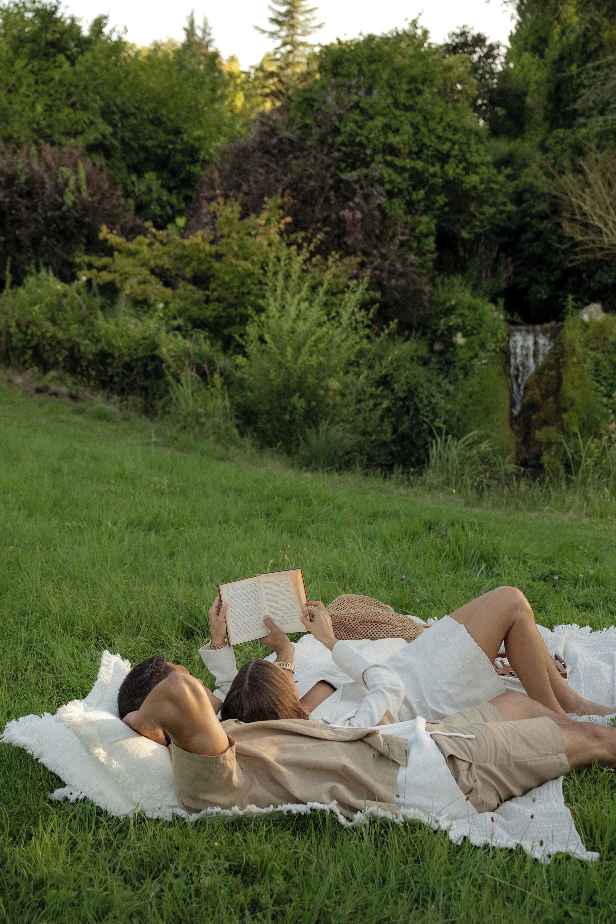 A person lying on a blanket in a grassy area with trees and a waterfall in the background.