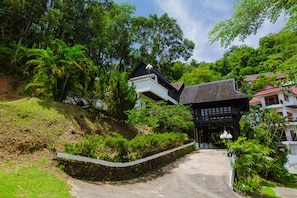 A traditional wooden house with a steeply sloped roof, surrounded by lush greenery and a stone wall.
