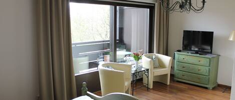 Une chambre d’hôtel avec un lit, une télévision, une commode verte, une chaise et une table sur laquelle est posé un vase de fleurs.