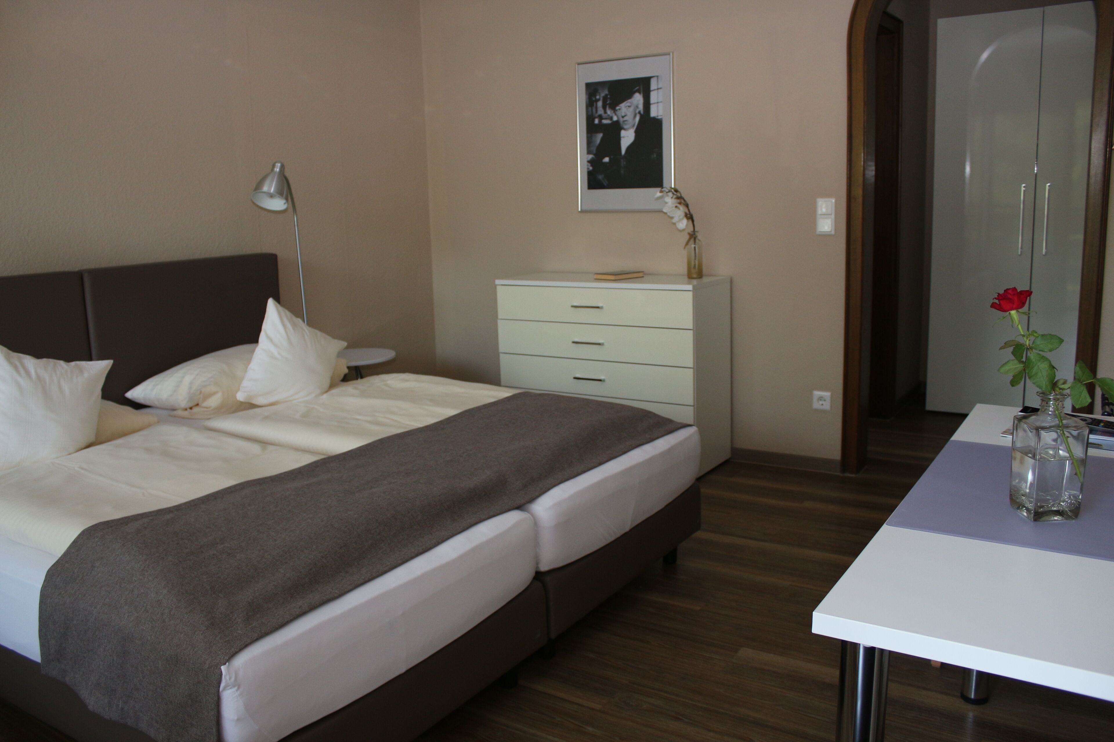 A bedroom with a bed, bedside table, lamp, dresser, and a framed picture on the wall.