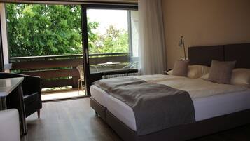 A modern hotel room with a large bed, a desk, and a balcony with a view of trees.