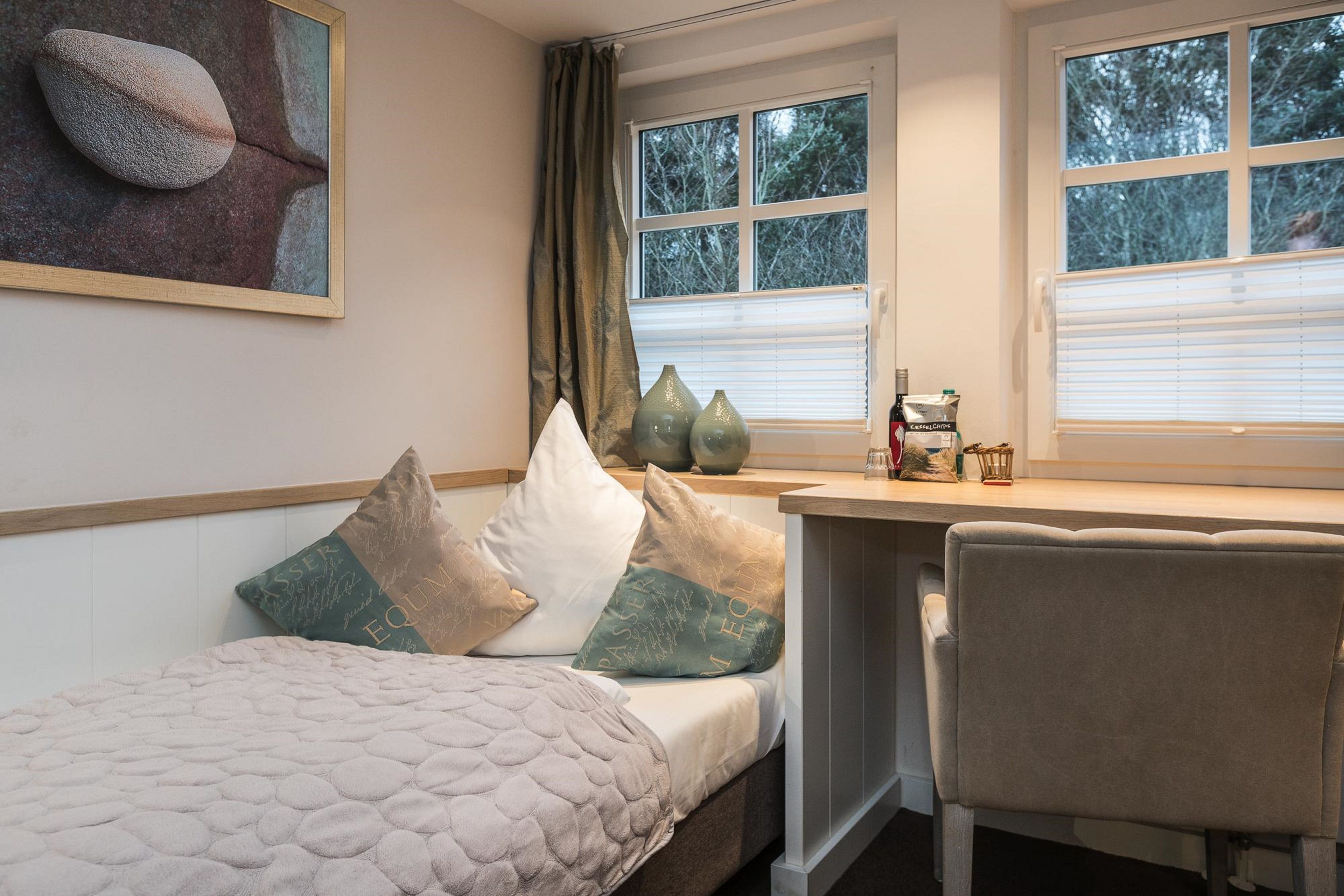 Een slaapkamer met een bed, een bureau en een stoel. Op het bureau staan drie vazen, er is een raam met zonwering en aan de muur hangt een schilderij.