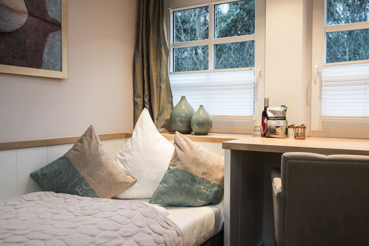 Un dormitorio con cama, almohadas, escritorio y ventana con persianas.